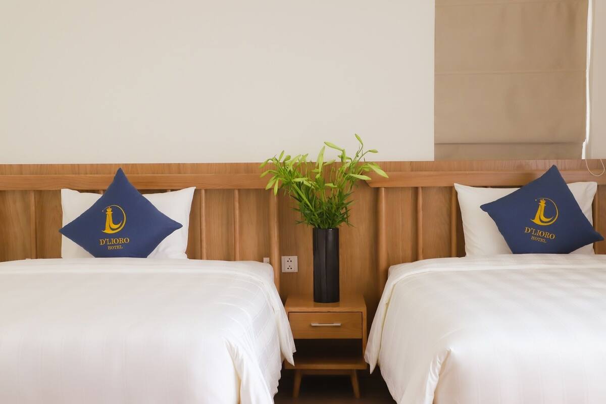 Une chambre d’hôtel avec deux lits, chacun agrémenté d’un oreiller orné du logo de l’hôtel. Une petite table de chevet en bois, dotée d’un tiroir, se trouve entre les deux lits.