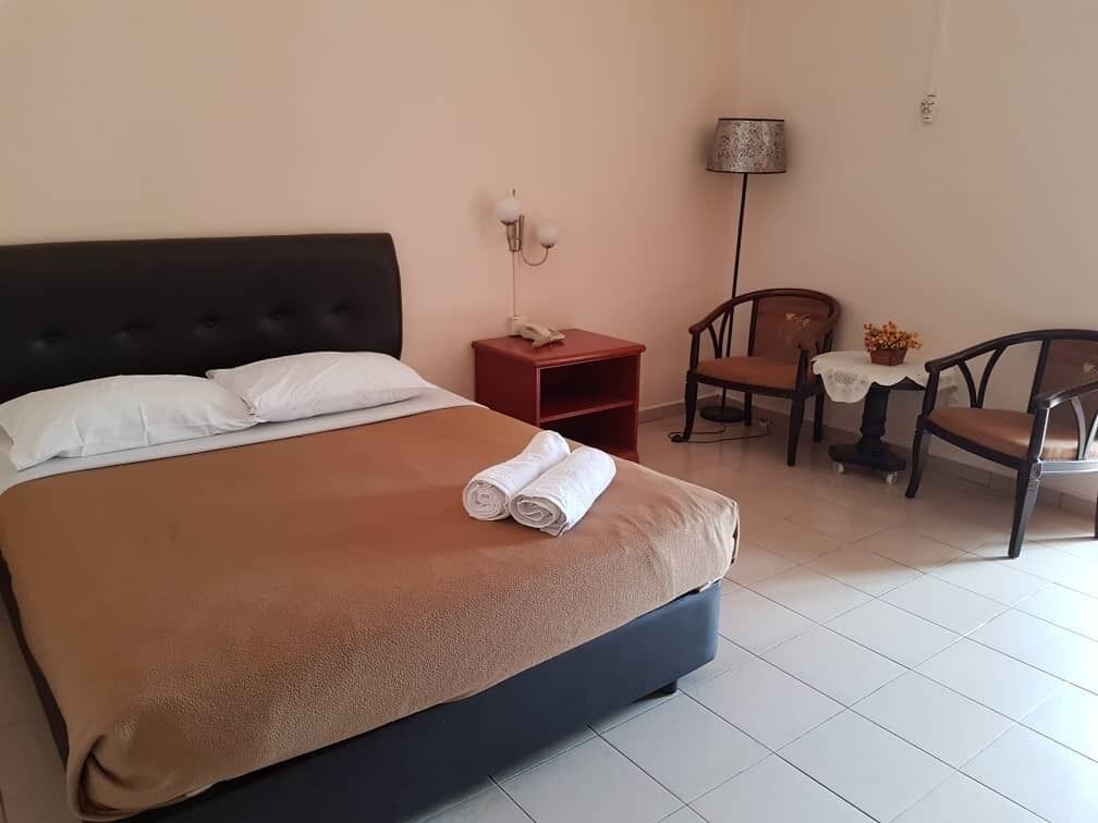 A neatly made bed with a brown cover, a bedside table with a lamp, and a chair with a table beside it.
