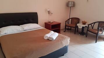 A neatly made bed with a brown cover, a bedside table with a lamp, and a chair with a table beside it.