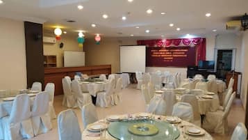 Un salón de banquetes con mesas redondas dispuestas para comer, sillas blancas y un escenario con fondo de cortina roja.