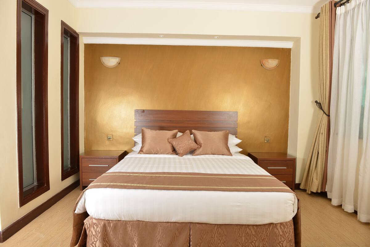 A hotel room with a large bed, two bedside tables, a wooden headboard, and a window with curtains.
