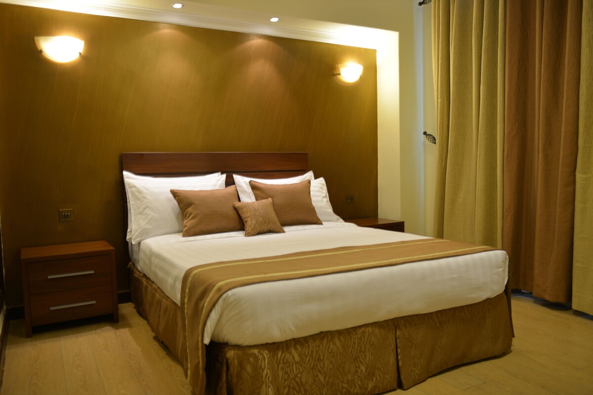 Habitación de hotel con una cama grande, cabecera de madera, dos mesitas de noche y una cortina en la ventana.