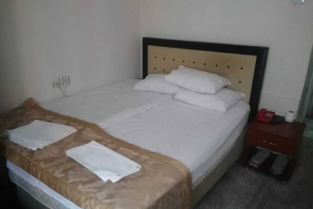A neatly made bed with white linens and a brown bedspread, a small bedside table with a red object, and a wall socket.