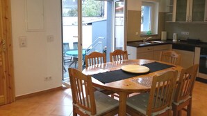 Un coin repas avec une table et des chaises en bois, une cuisine équipée d’un évier et de placards, et une porte-fenêtre coulissante donnant sur une terrasse extérieure.