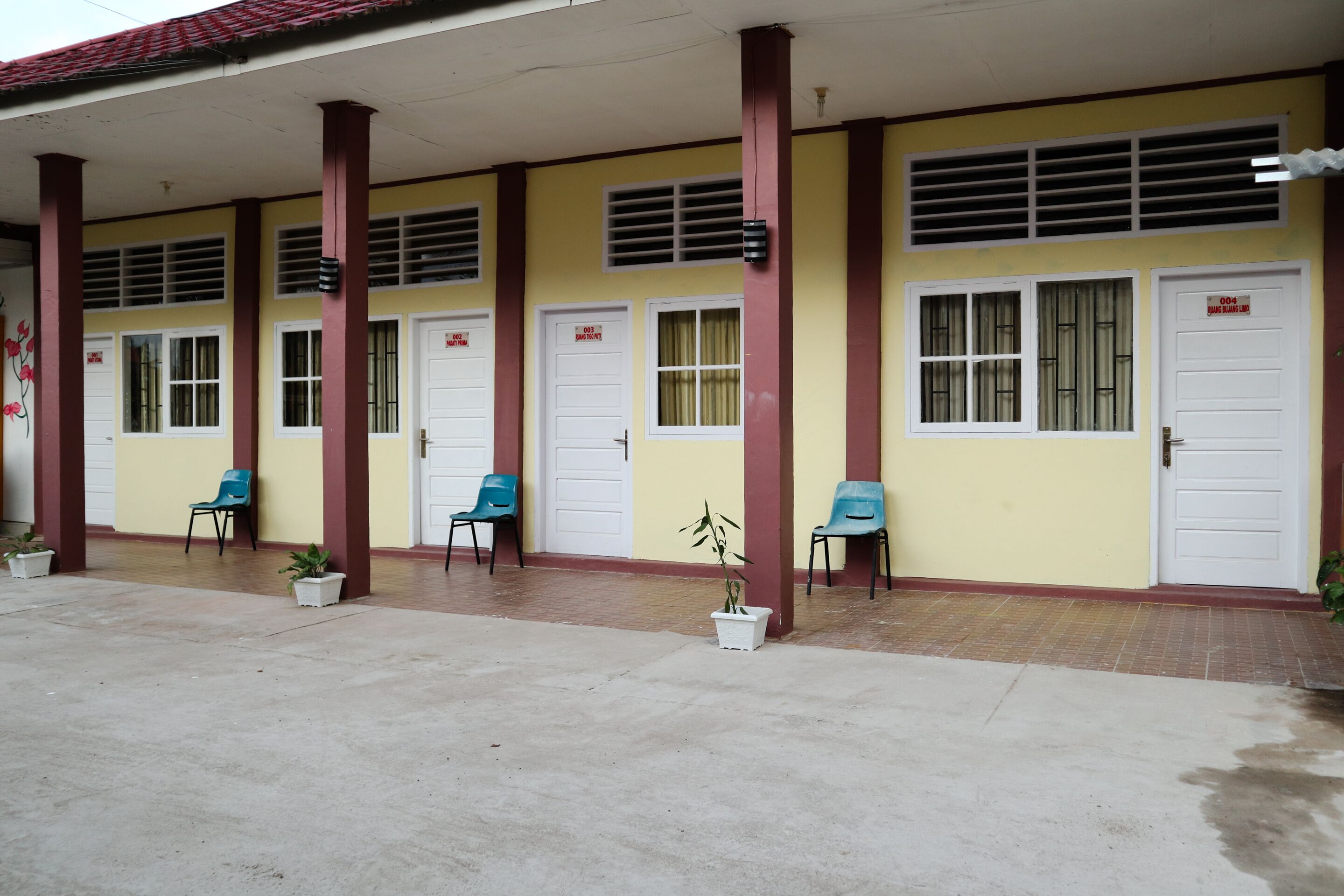 A building with a yellow facade, white doors, and red pillars. There are potted plants and blue chairs outside.