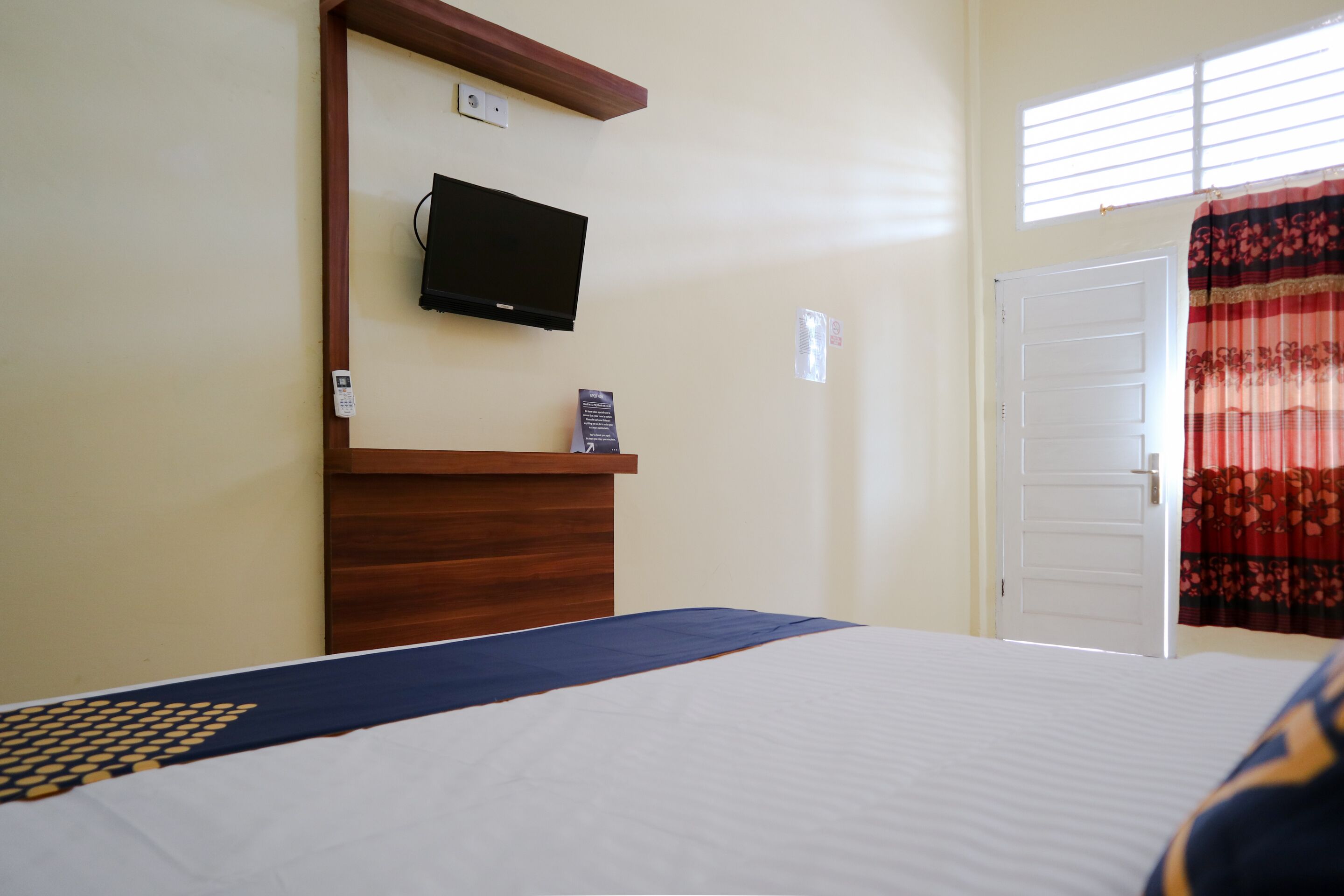 Une chambre d’hôtel avec un lit, une tête de lit en bois, un téléviseur à écran plat fixé au mur, une porte et une fenêtre avec des rideaux.