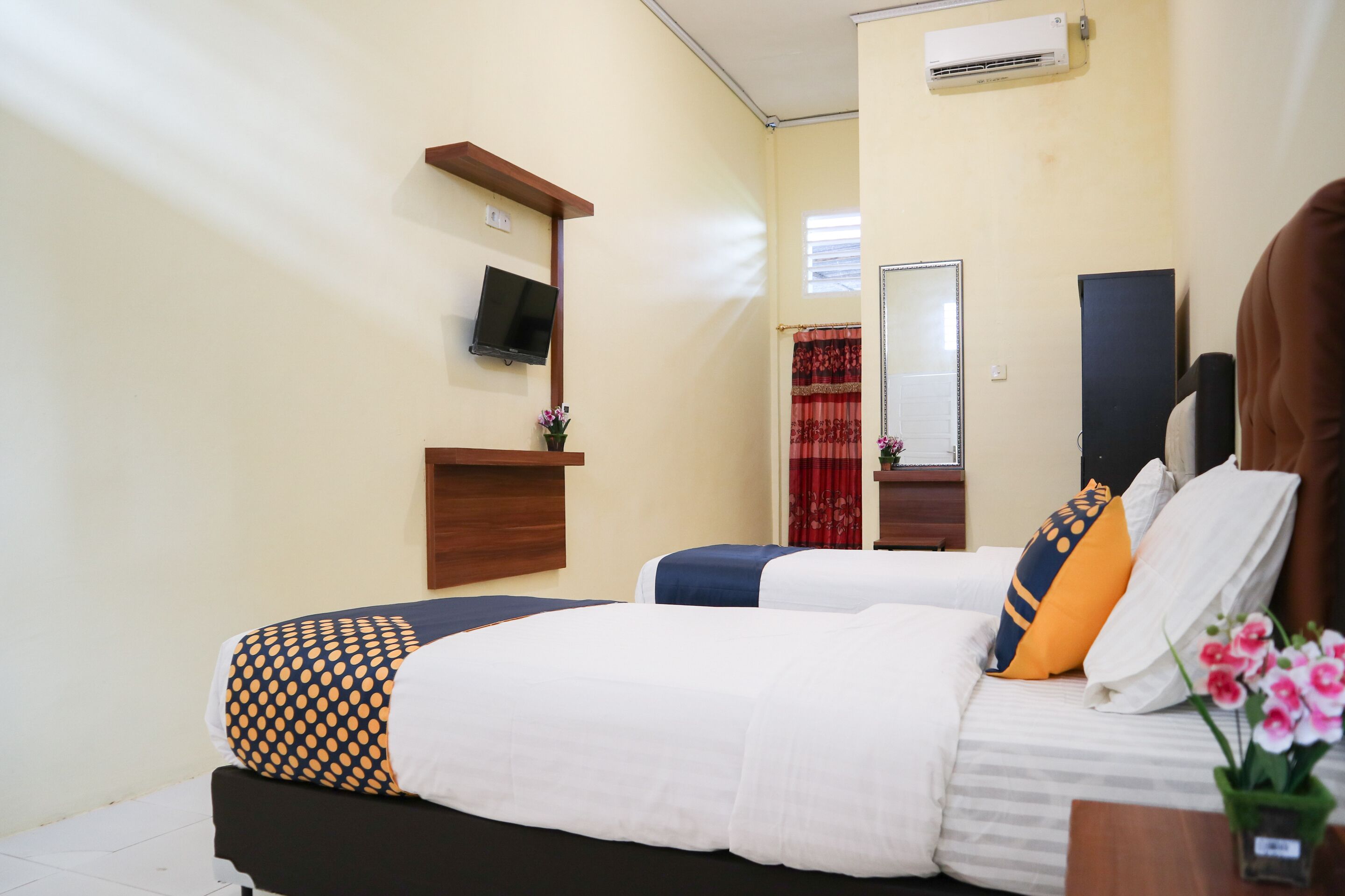 A hotel room with two beds, a TV mounted on the wall, a small table with a vase of flowers, and an air conditioning unit.