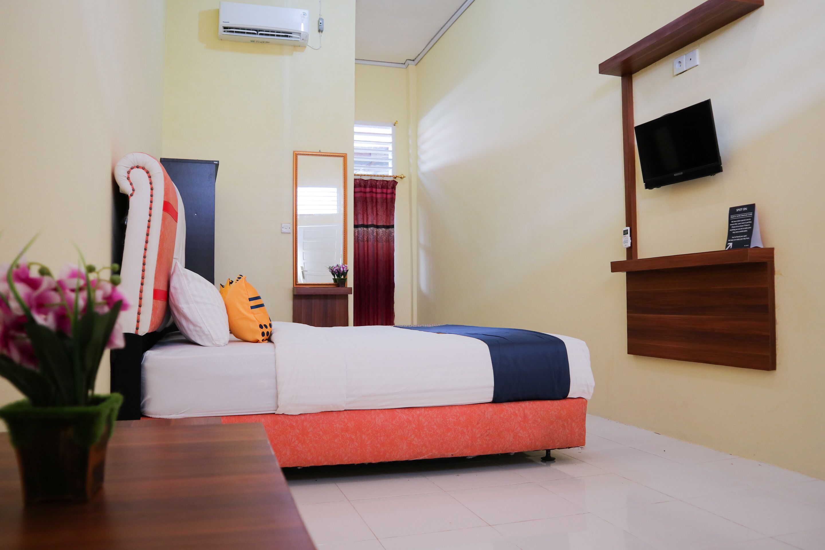 A hotel room with a bed, a TV mounted on the wall, a small table with a vase of flowers, and a window with curtains.