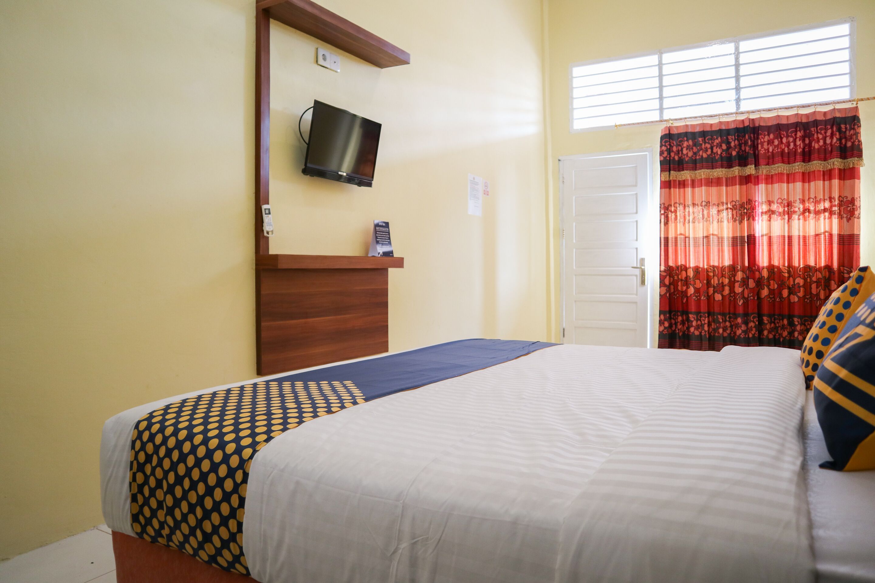 A hotel room with a bed, a TV mounted on the wall, a door, and a window with curtains.