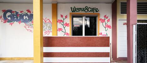 A reception area with a wooden bench, a wall with floral graffiti, and a sign reading "Wisma Cafe".