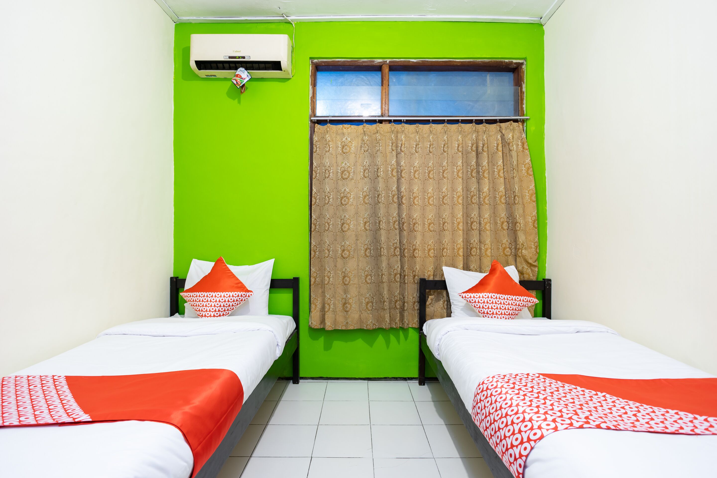Una habitación con dos camas, paredes verdes, una ventana con cortinas y un aire acondicionado.