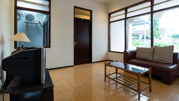 A room with a TV on a stand, a glass table, a brown leather sofa, and a window with a view of a building and greenery. - OYO 2454 Wanawiyata Homestay  (Depok)
