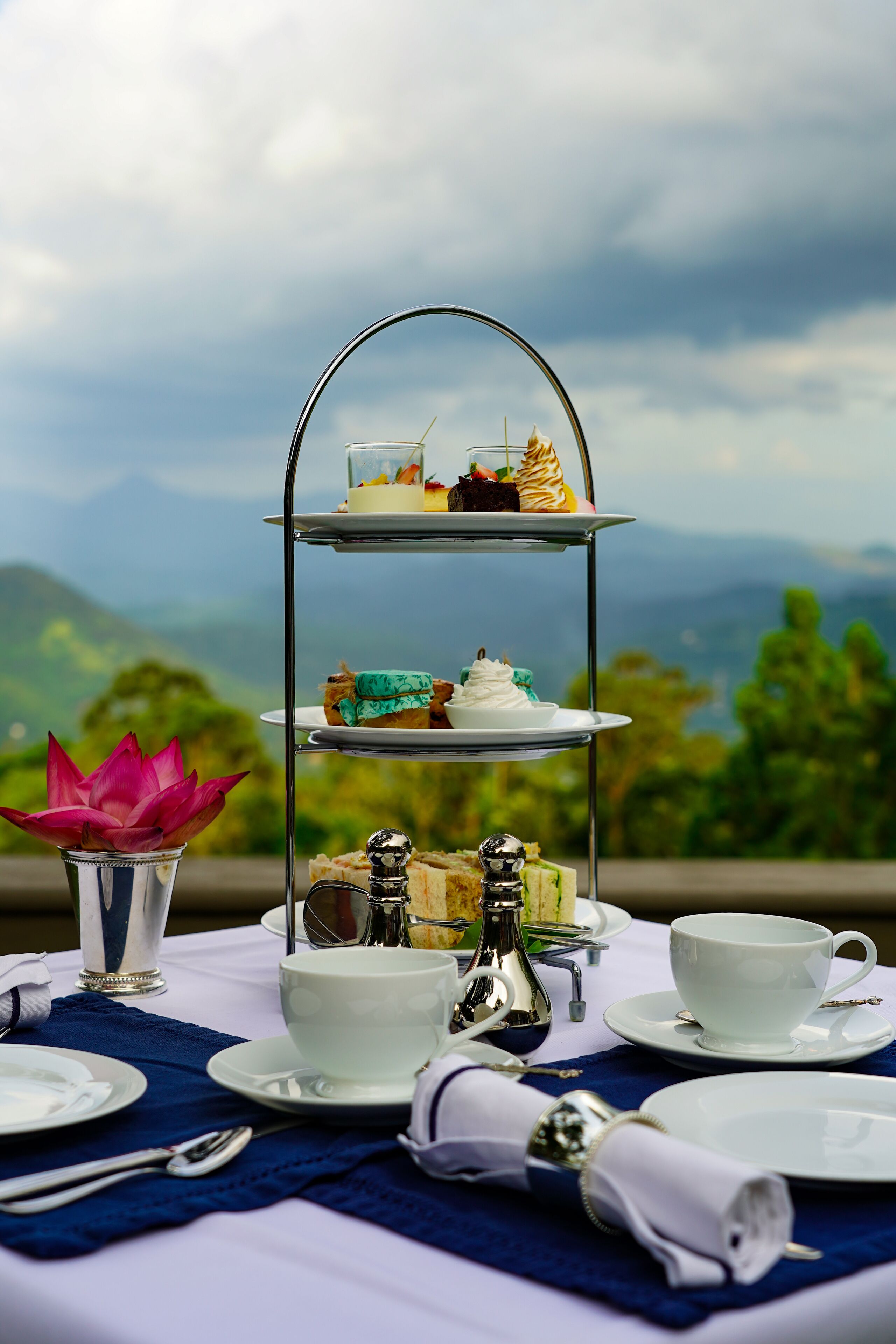 A three-tiered dessert stand with various pastries, a cup of tea, and a decorative flower arrangement on a table set against a scenic mountain backdrop.