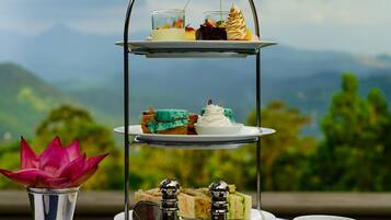 A three-tiered dessert stand with various pastries, a cup of tea, and a decorative flower arrangement on a table set against a scenic mountain backdrop.