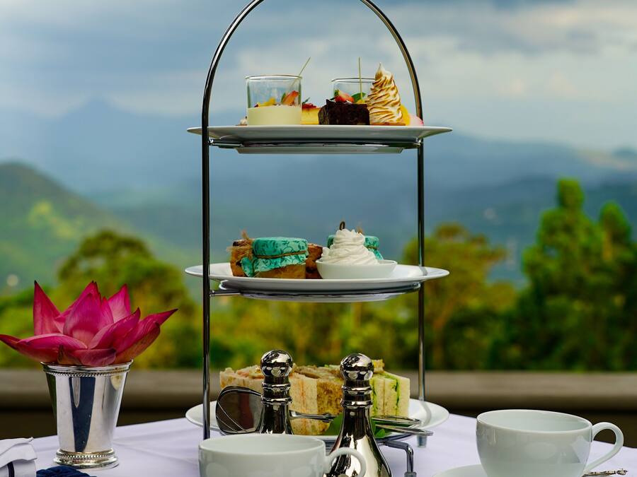 A three-tiered dessert stand with various pastries, a cup of tea, and a decorative flower arrangement on a table set against a scenic mountain backdrop.