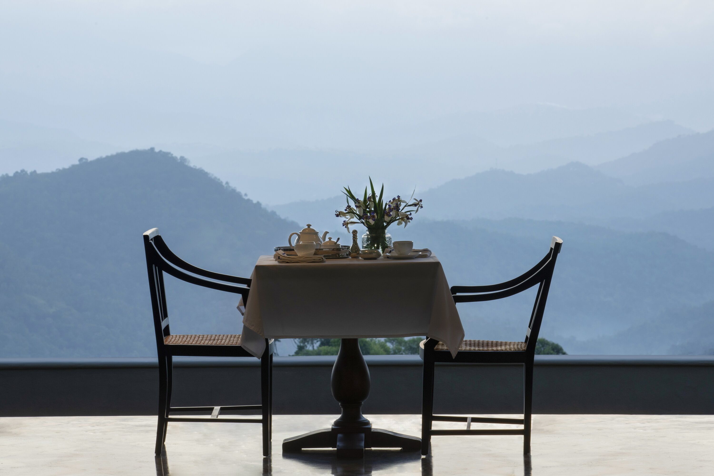 A table set with a vase of flowers, teapot, cups, and saucers, with a view of mountains in the background.