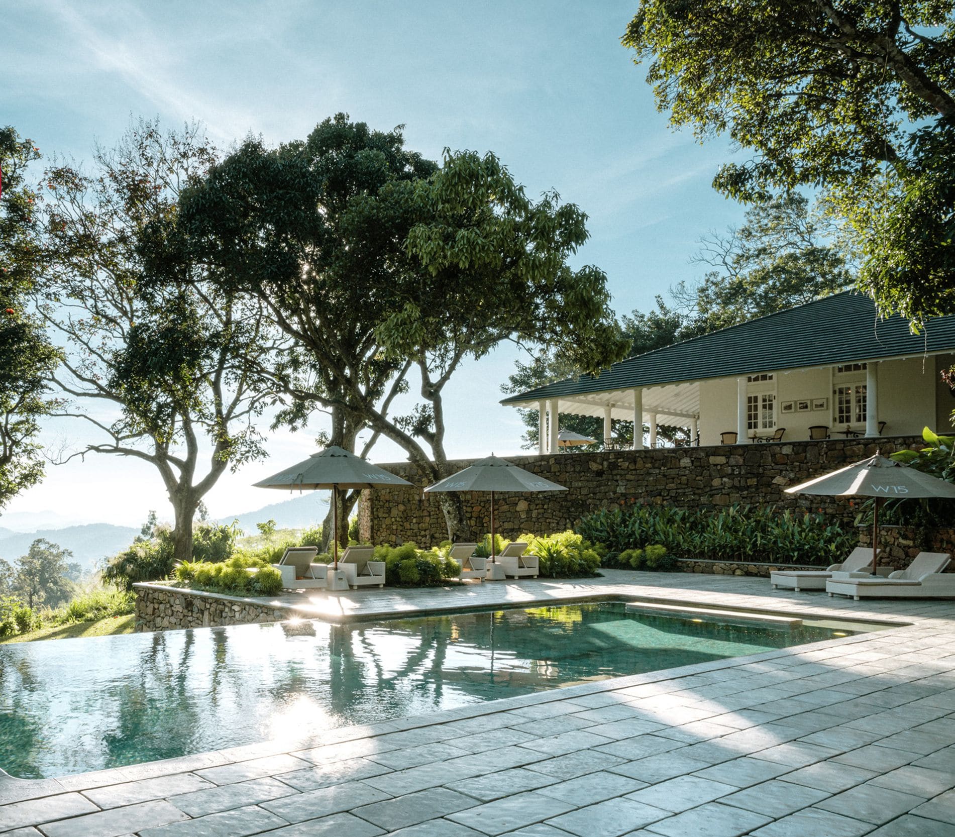 A luxurious outdoor pool area with lounge chairs and umbrellas, surrounded by trees and a stone wall, with a view of the mountains in the background.