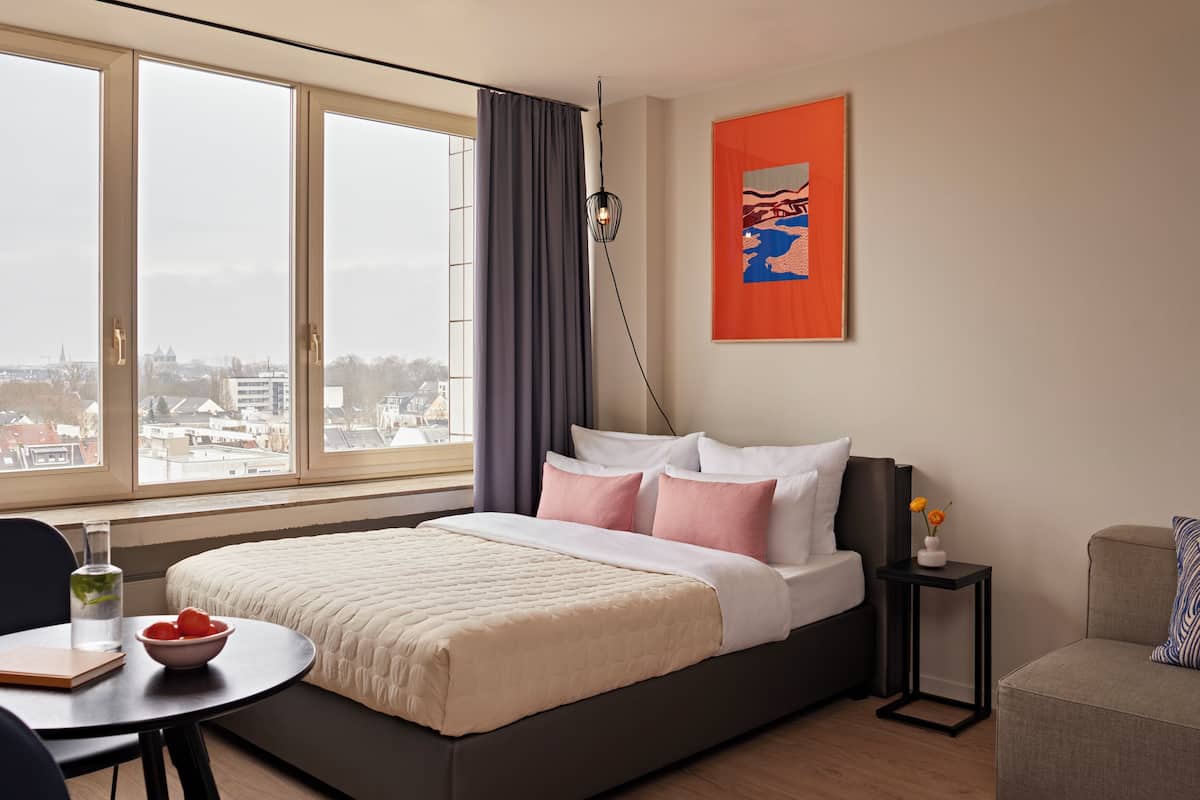 A hotel room with a bed, a desk, a chair, a table, a sofa, a window with curtains, a lamp, and a painting on the wall.