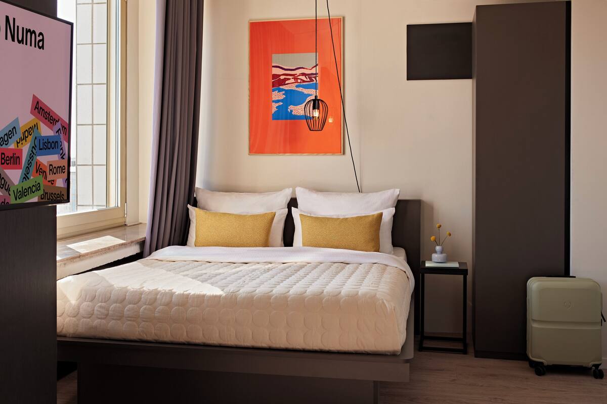 A hotel room with a bed, a nightstand, a lamp, a small table with a vase, and a wardrobe.