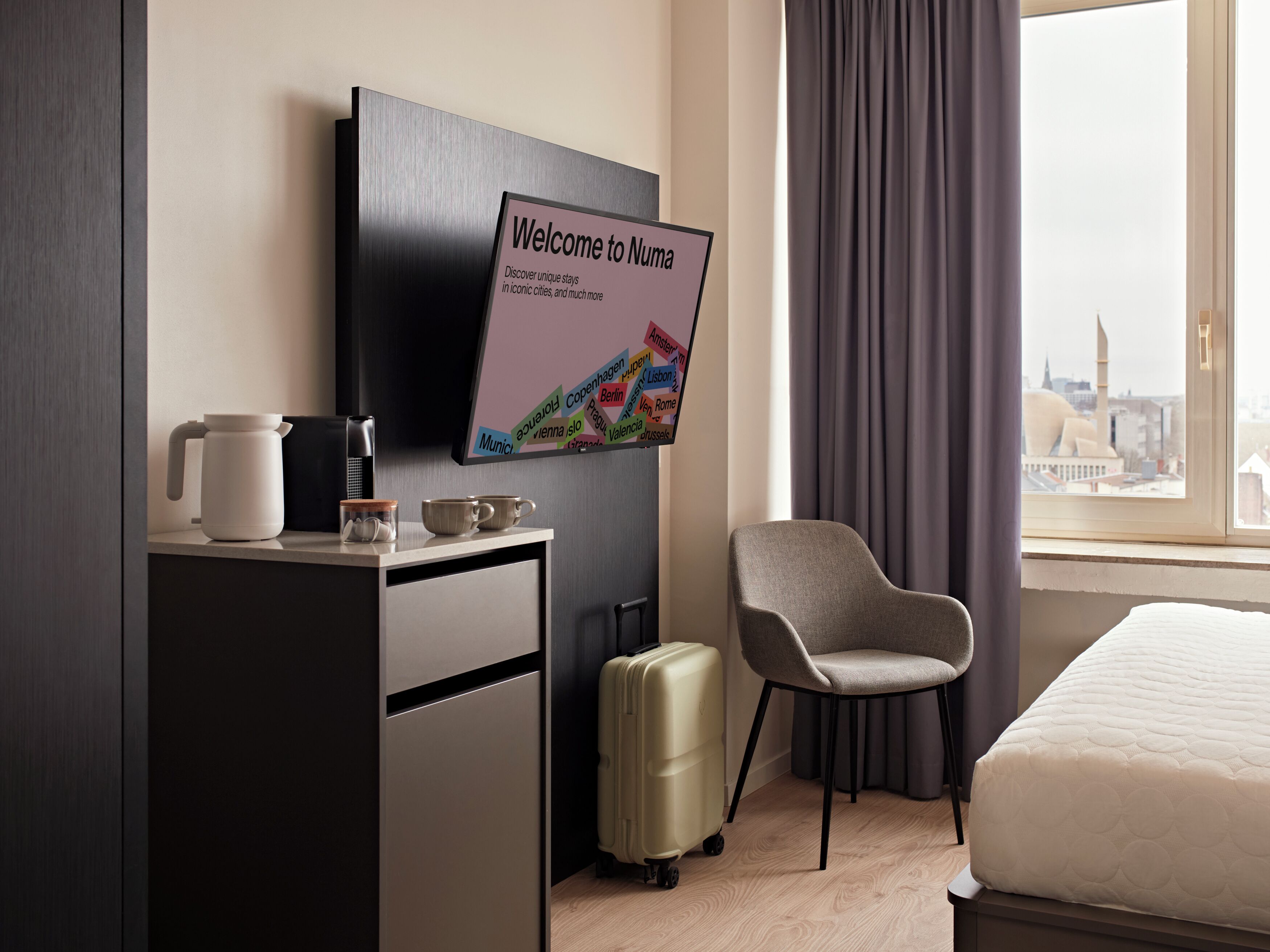 A hotel room with a bed, a chair, a desk with a coffee maker, a suitcase, a TV displaying a welcome message, and a window with a city view.