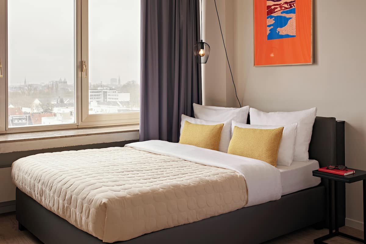 A modern hotel room with a large bed, a nightstand, a framed picture, and a view of the city.