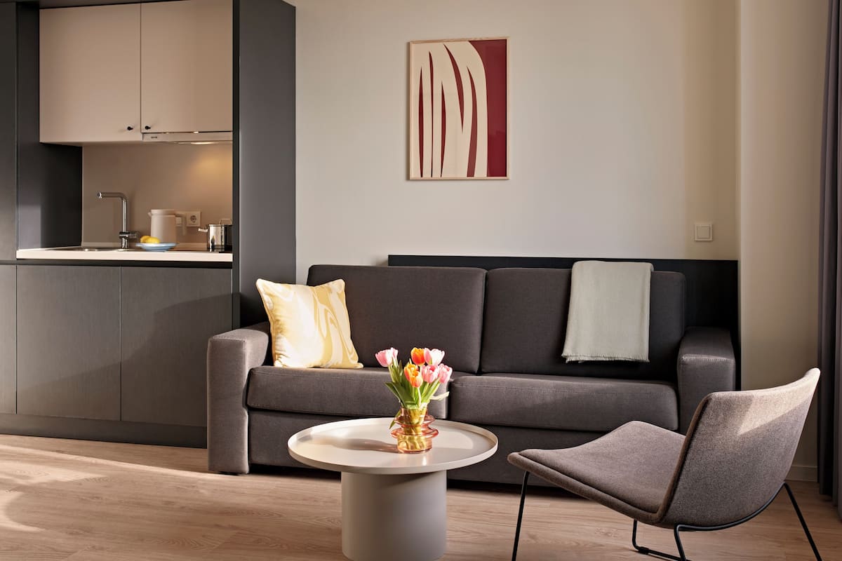 A modern living room with a sofa, a coffee table, and a chair. There is a kitchen area with cabinets and a sink.