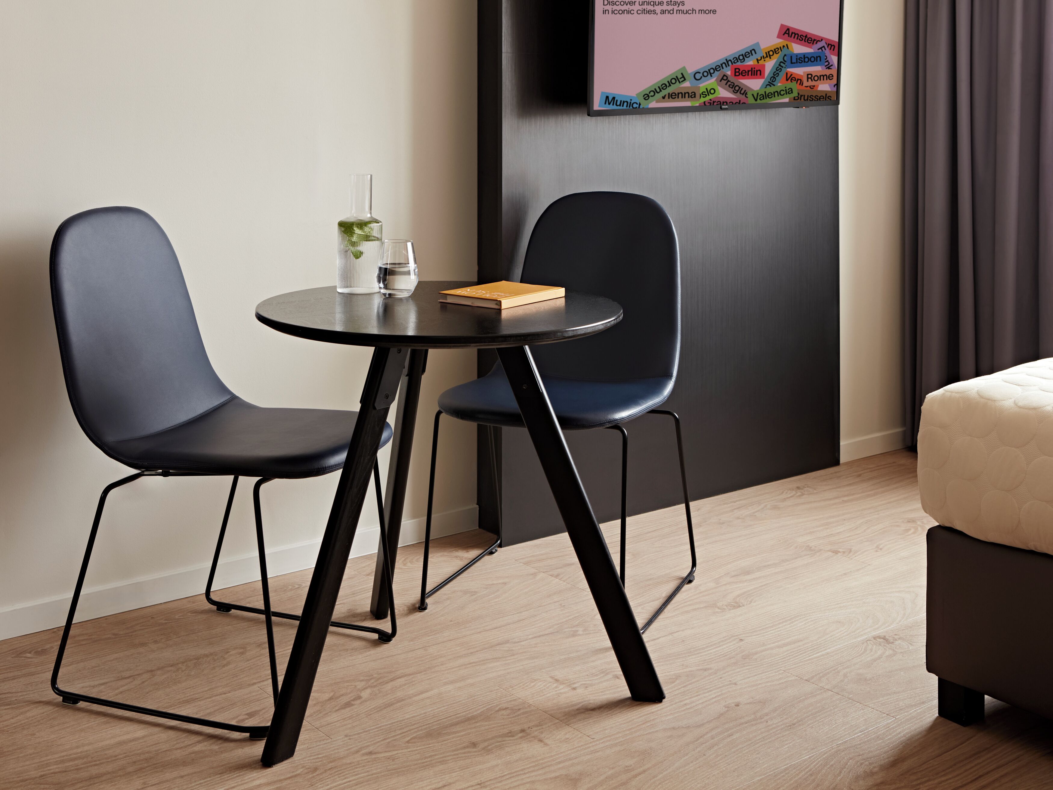 Ein modernes Hotelzimmer mit Holzboden, zwei schwarzen Stühlen um einen kleinen runden Tisch, einem Bett mit gestepptem Kopfteil und einem an der Wand befestigten Fernseher, der Reiseziele zeigt.