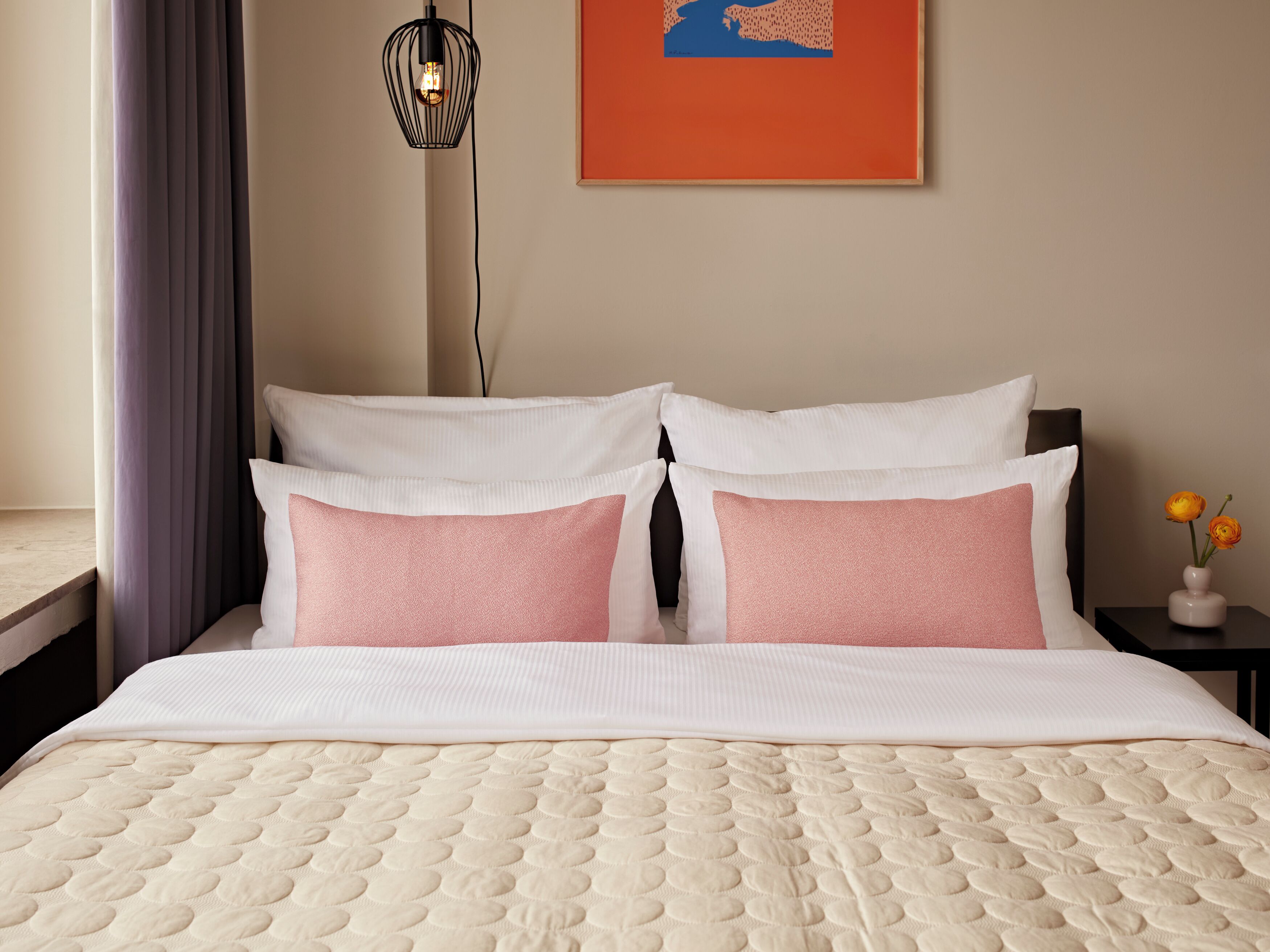 Una cama bien tendida con cubrecolcha acolchado, dos almohadas, una mesita de noche con un florero y una lámpara fijada a la pared.