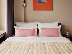 Cama bem arrumada com cobertura acolchoada, dois travesseiros, mesa de cabeceira com um vaso de flores e luminária fixada na parede.