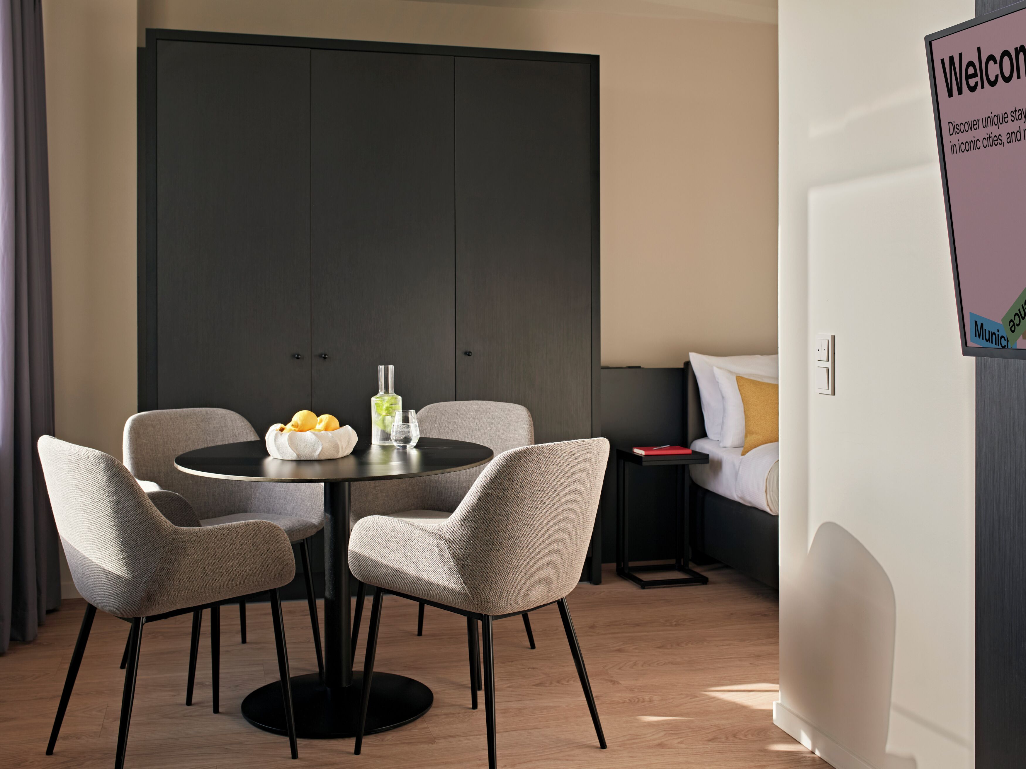 A modern hotel room with a round dining table, four chairs, a minibar, a bed with white and mustard pillows, and a welcome sign.