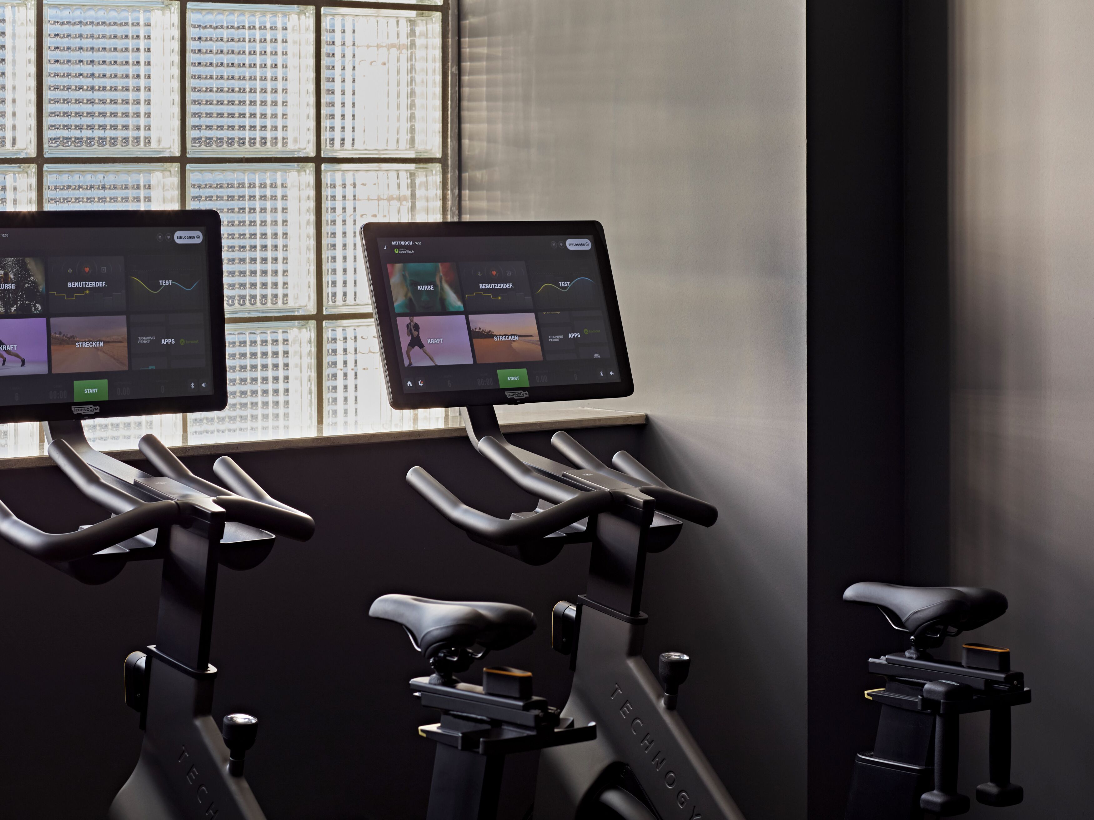 Tre motionscyklar med digitala displayer, ett fönster med persienner och en väggmonterad kontrollpanel.