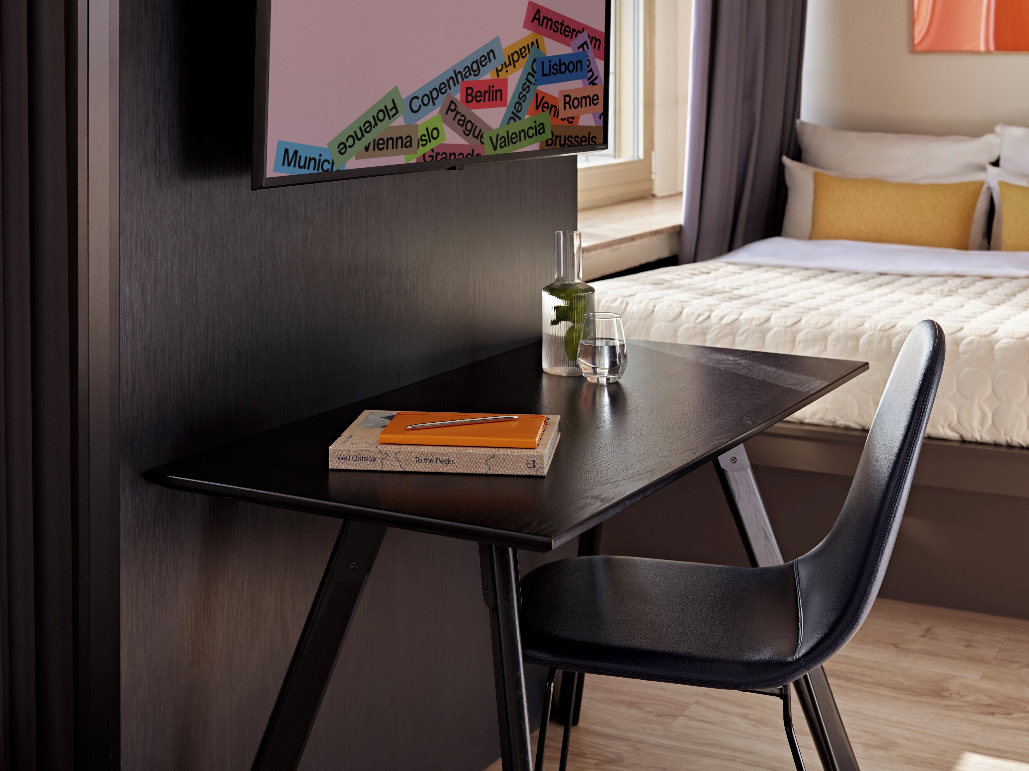 Een moderne hotelkamer met een donker houten bureau, een stoel, een bed met witte beddengoed en een aan de muur bevestigde televisie waarop verschillende stadsnamen te zien zijn.
