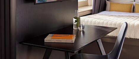 Een moderne hotelkamer met een donker houten bureau, een stoel, een bed met witte beddengoed en een aan de muur bevestigde televisie waarop verschillende stadsnamen te zien zijn.