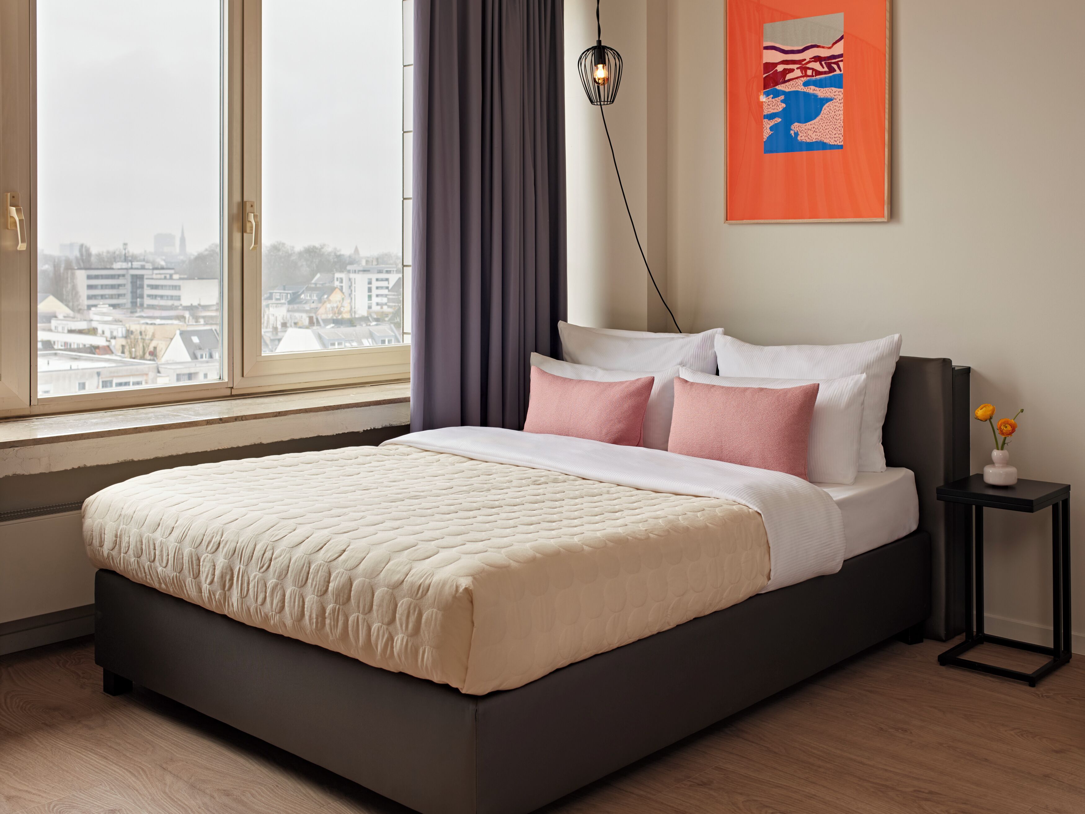 A modern bedroom with a large bed, a city view, a framed artwork, and a bedside table with flowers.