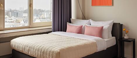 A modern bedroom with a large bed, a city view, a framed artwork, and a bedside table with flowers.
