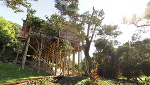 A wooden structure with a balcony, surrounded by trees and greenery. - Campismo Ninho 28 (Ponta d'Ouro)