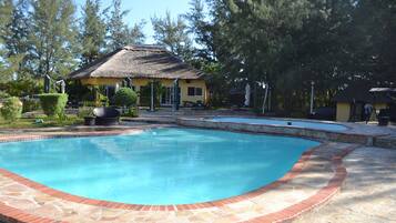 A swimming pool with a brick border, surrounded by a patio area with a thatched-roof building and lush greenery.