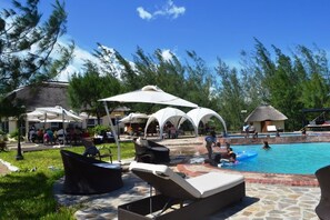 Área de piscina com espreguiçadeiras e guarda-sóis, cercada por árvores e uma construção com telhado de palha.