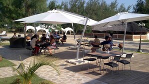 An outdoor patio area with tables and chairs under white umbrellas, surrounded by trees and a pool.