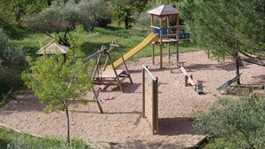 A playground with a slide, swings, and climbing structures surrounded by trees and a house in the background.