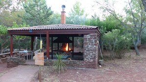 A rustic outdoor cooking area with a brick chimney, surrounded by trees and a stone wall.