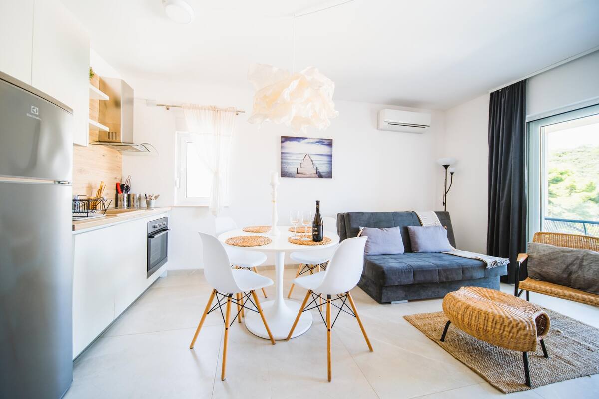 A modern kitchen with white cabinets, a dining table with white chairs, a gray sofa, and a wicker ottoman.