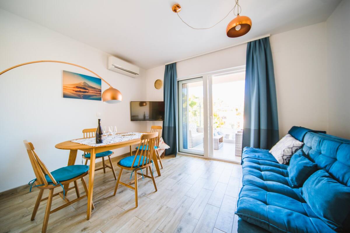 A modern living room with a blue sofa, wooden dining table with chairs, a TV, and a large window with curtains.