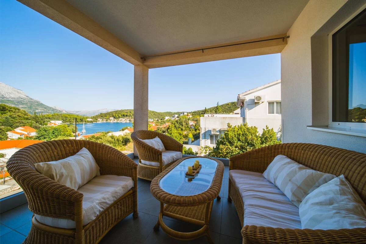 A balcony with wicker furniture, a glass table, and a view of a mountainous landscape.