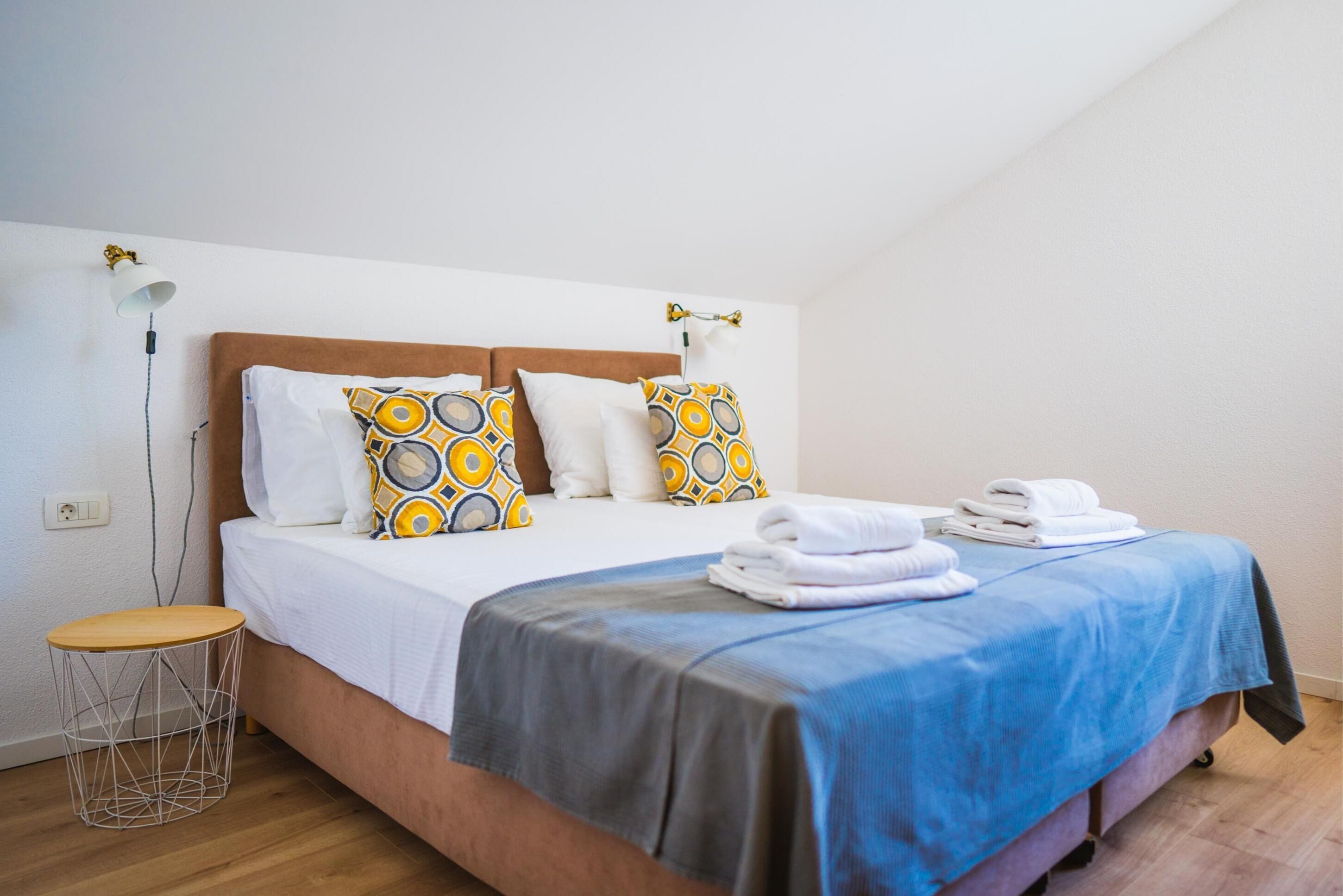 A neatly made bed with patterned pillows, a blue bedspread, and folded towels on the side.