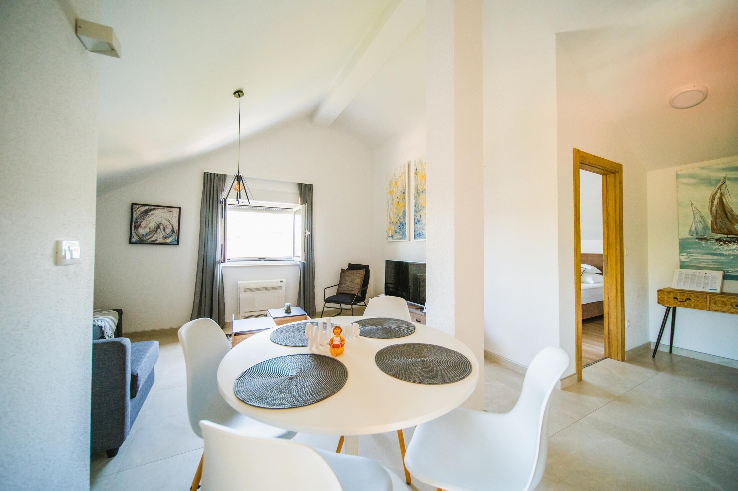 Un espace repas moderne avec une table et des chaises blanches, un luminaire suspendu et une vue sur une chambre où se trouve un tableau.