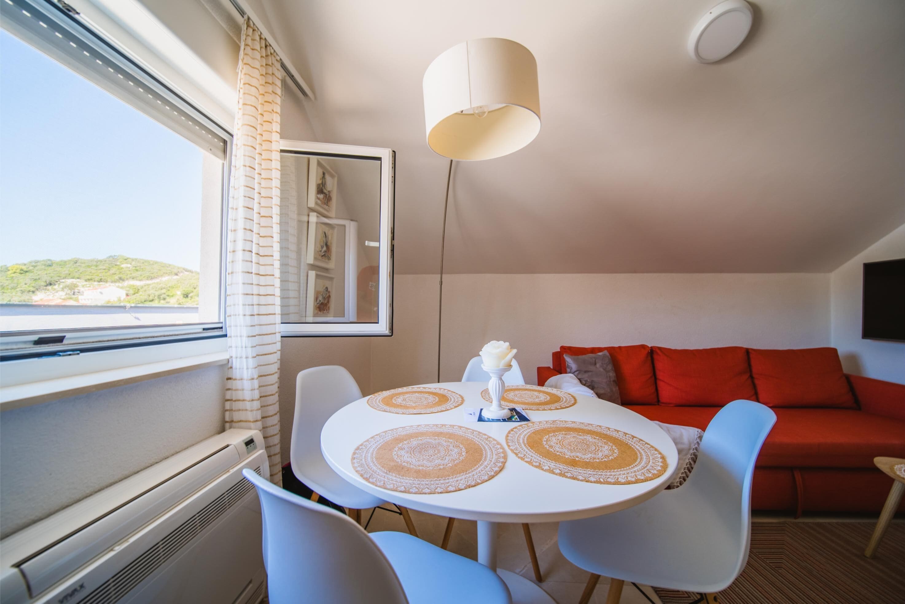 A dining area with a round table, white chairs, and a white tablecloth. There is a red sofa and a window with a view of a green landscape.