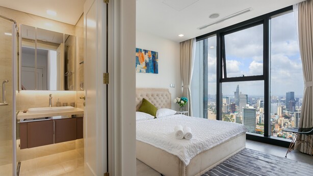 A hotel room with a large bed, a city view, a bathroom with a mirror and sink, and a window with curtains. - Stephen's Riverside Apartments (Ho Chi Minh City)