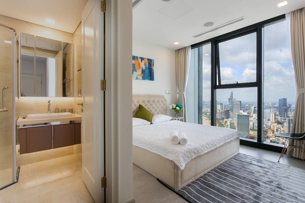A hotel room with a large bed, a city view, a bathroom with a mirror and sink, and a window with curtains.