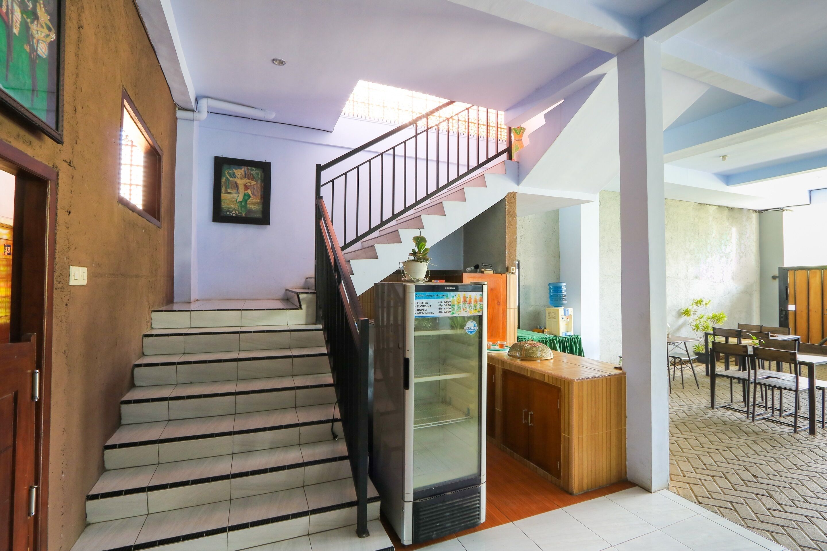 Un escalier menant à un deuxième étage, un réfrigérateur, un coin repas avec des chaises et des tables, et un mur orné d’un tableau encadré.